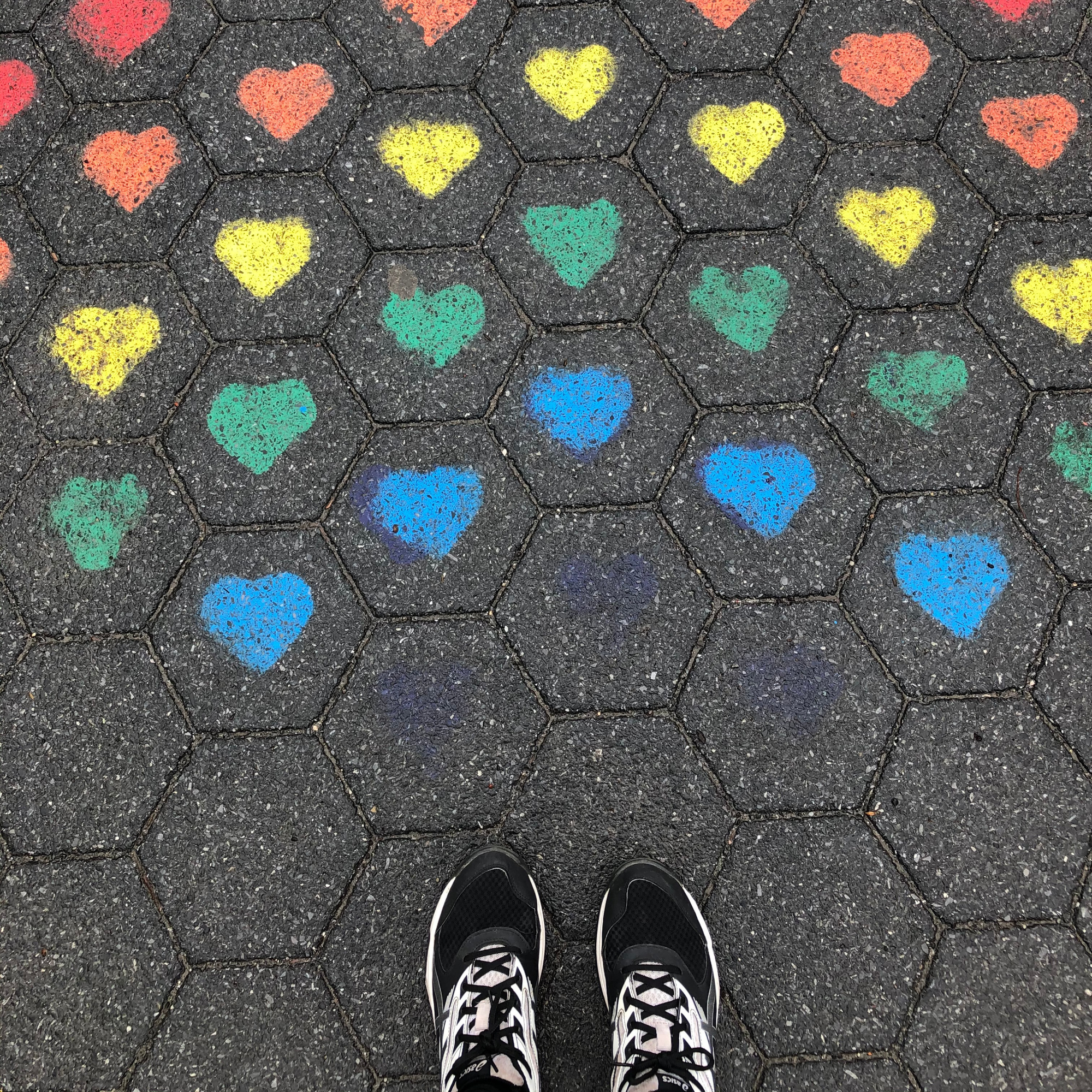 looking down at a person's sneakers standing in front of a series of colorful charcoal hearts drawn on a sidewalk