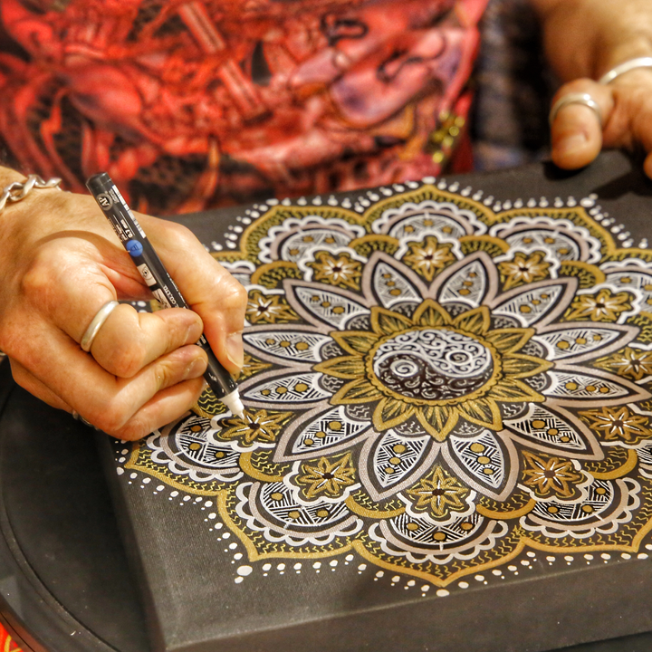 A closeup of a woman's hands drawing a gold and white mandala design in pen.